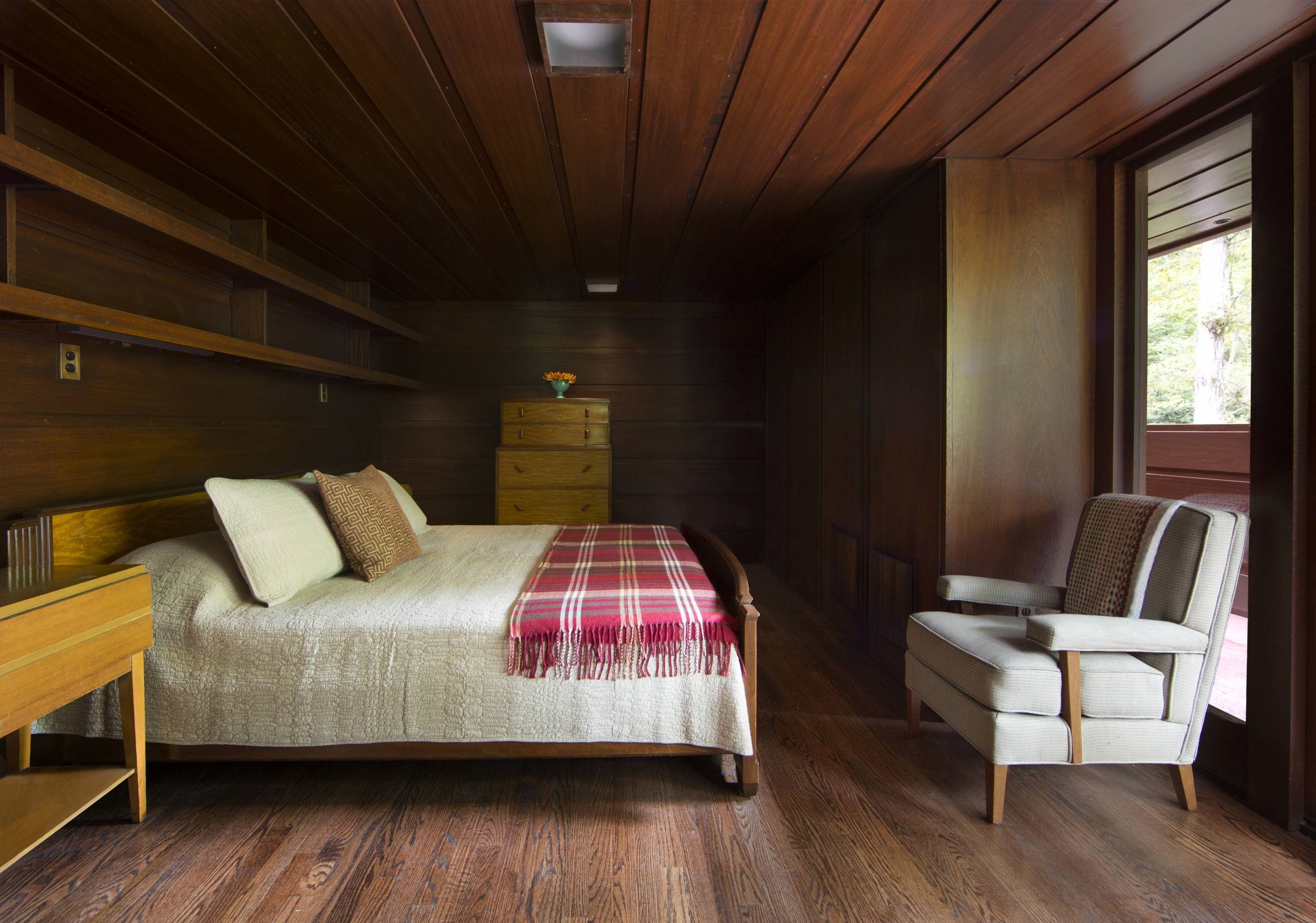 Bedroom with wooden paneling, double bed, quilt, plaid blanket, armchair, drawers, shelves.
