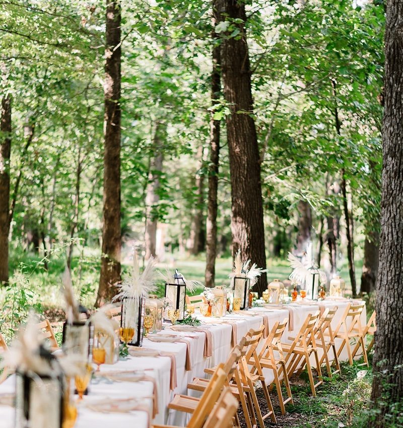 Elegantly set outdoor table with flowers, lanterns, and glassware in a forest setting.