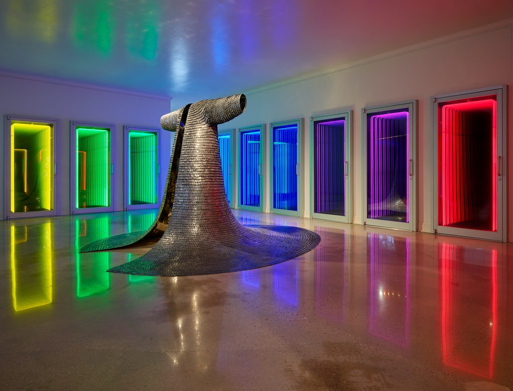 Metallic cloak sculpture in gallery with neon-lit rectangular frames in rainbow colors.