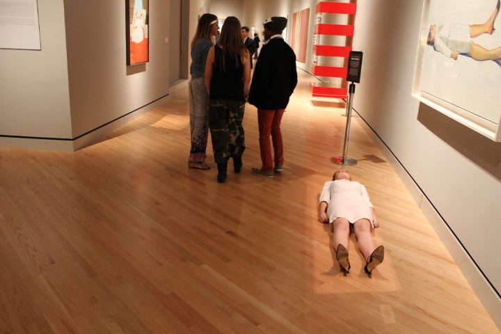 Art gallery with people observing, woman lies on floor, paintings displayed on walls.