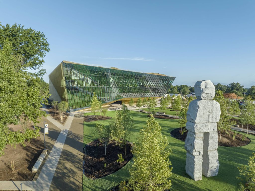 Modern glass building with angled walls beside a landscaped area with trees and stone sculpture.