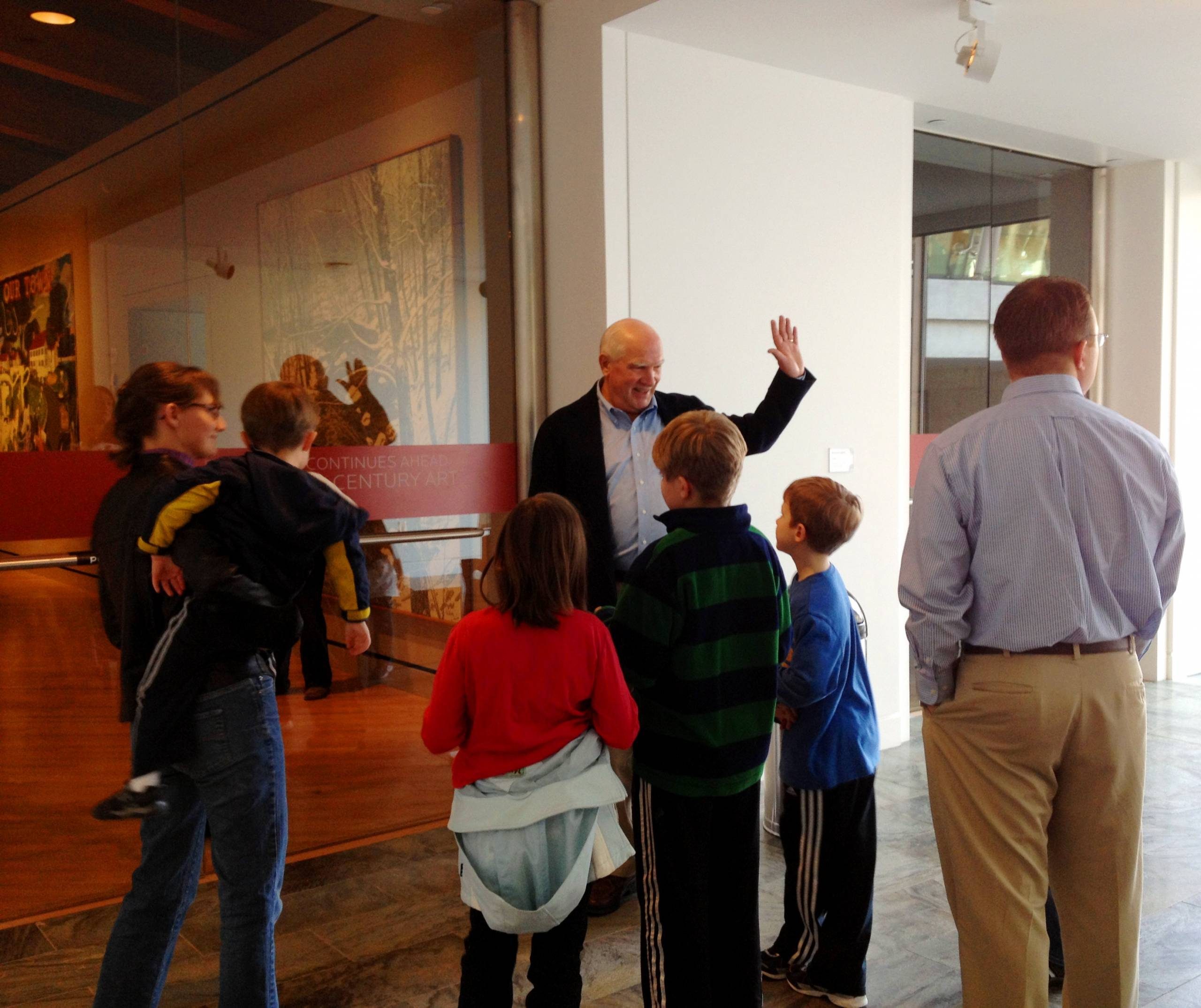 Group of people, including children, gathered indoors, interacting with an older man near artwork.