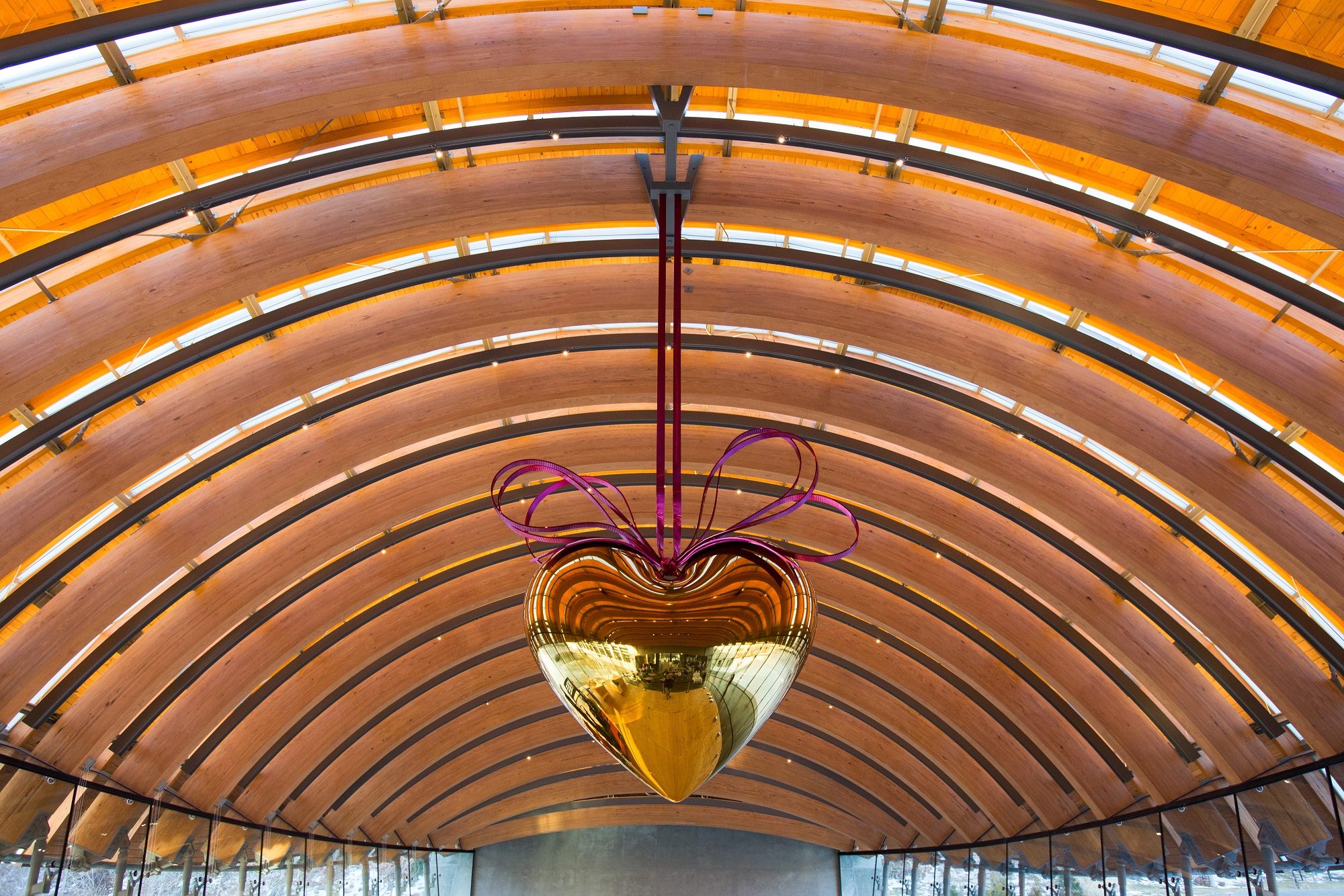 Gold heart sculpture with purple ribbons hangs under arched wooden roof with glass windows.