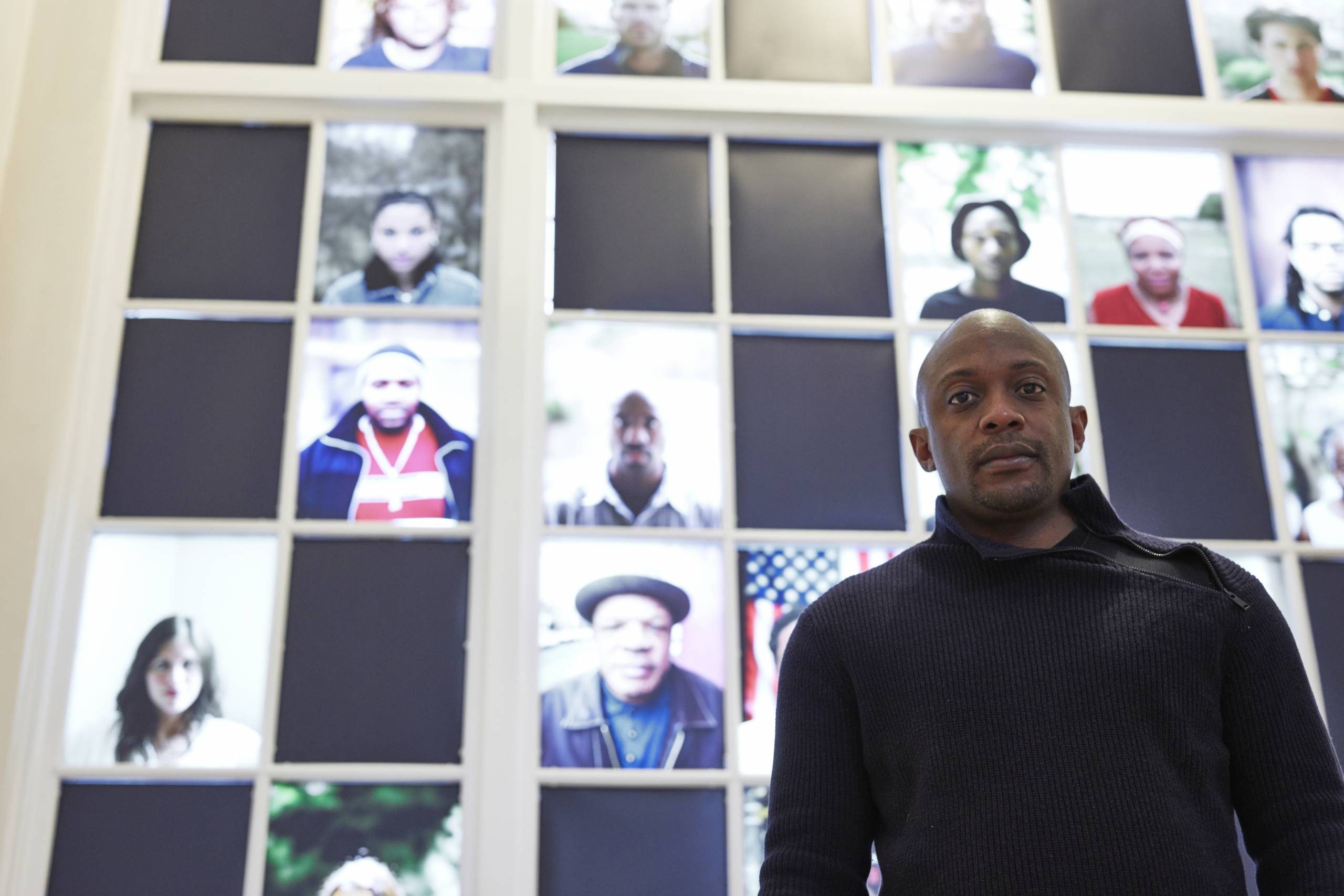 Man indoors in front of a window with a grid of illuminated portraits.