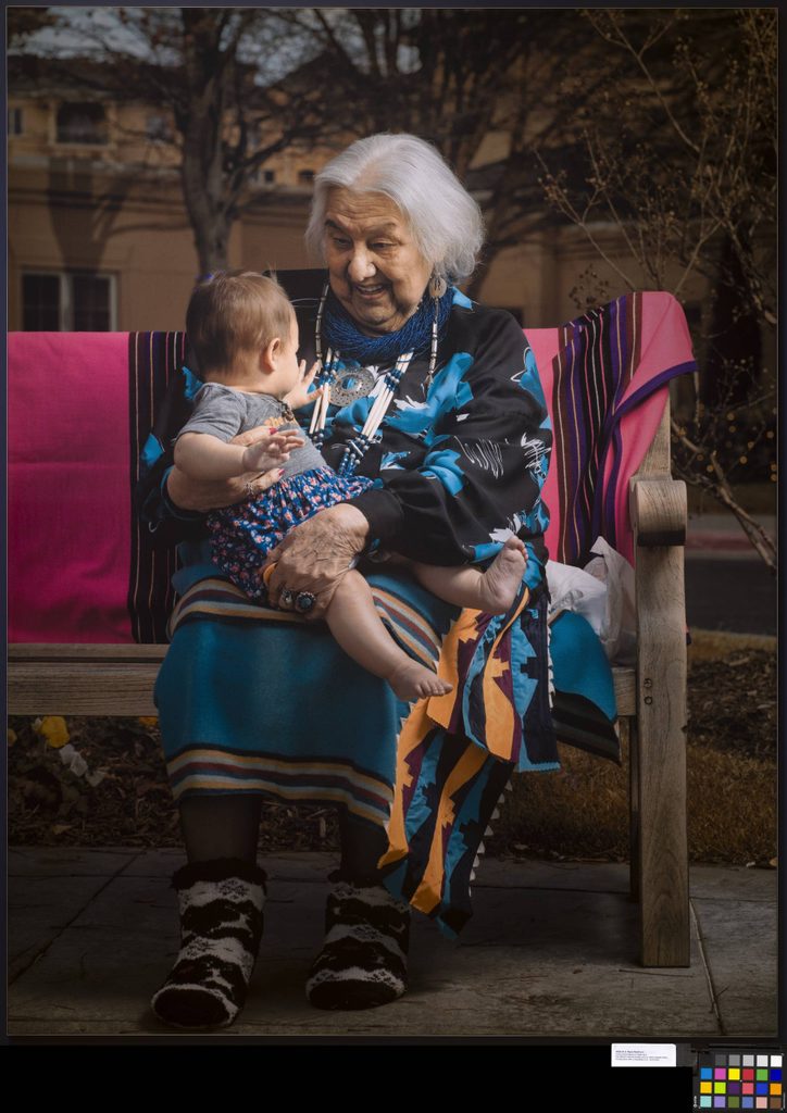 Elderly woman with baby on bench, colorful textiles, patterned attire, outdoor setting.