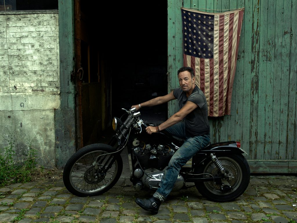 Man sitting on motorcycle in front of an old building with an old United States flag hanging on it