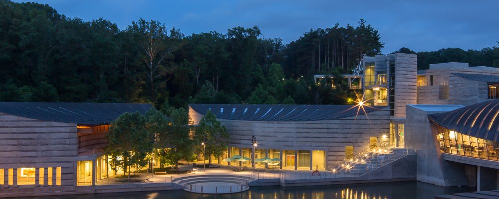 Modern architectural complex with curved roofs, large windows, forest, and water reflections at dusk.