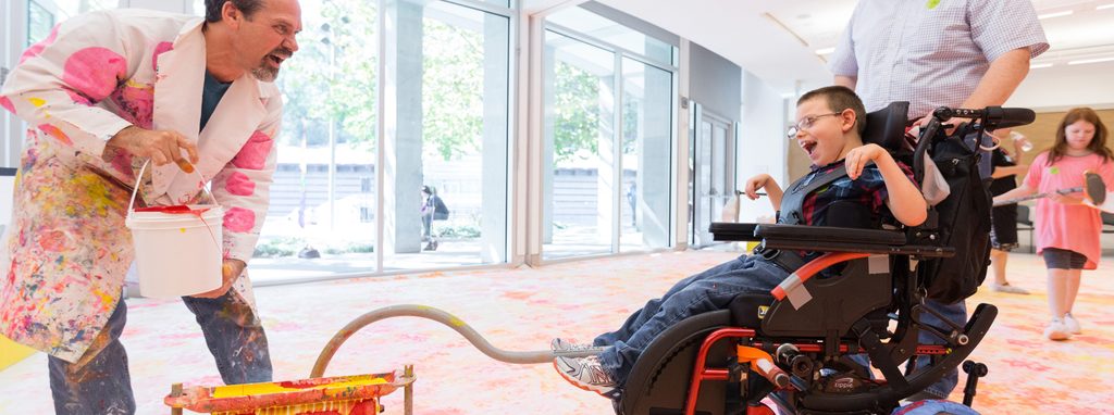 Man in paint-splattered coat engages child in wheelchair during art activity in bright room.