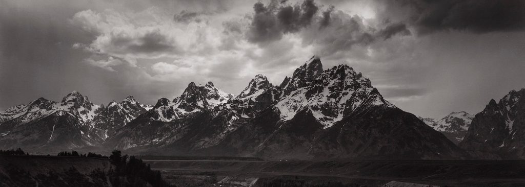 Rugged mountain range with snow-capped peaks, cloudy sky, wooded area, and valley in foreground.