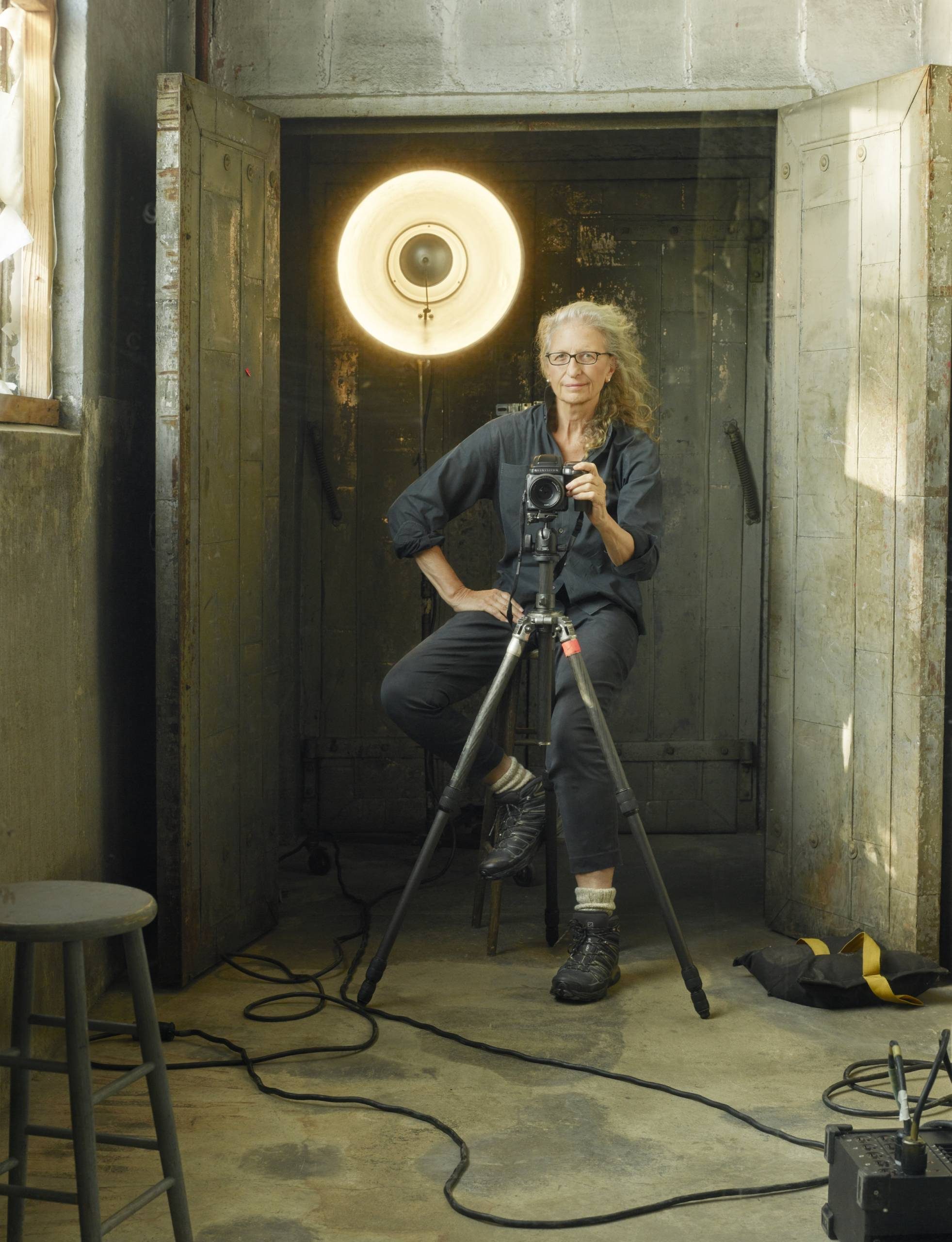 Women sits on stool in doorway with larger light behind her and camera on tripod in front of her.