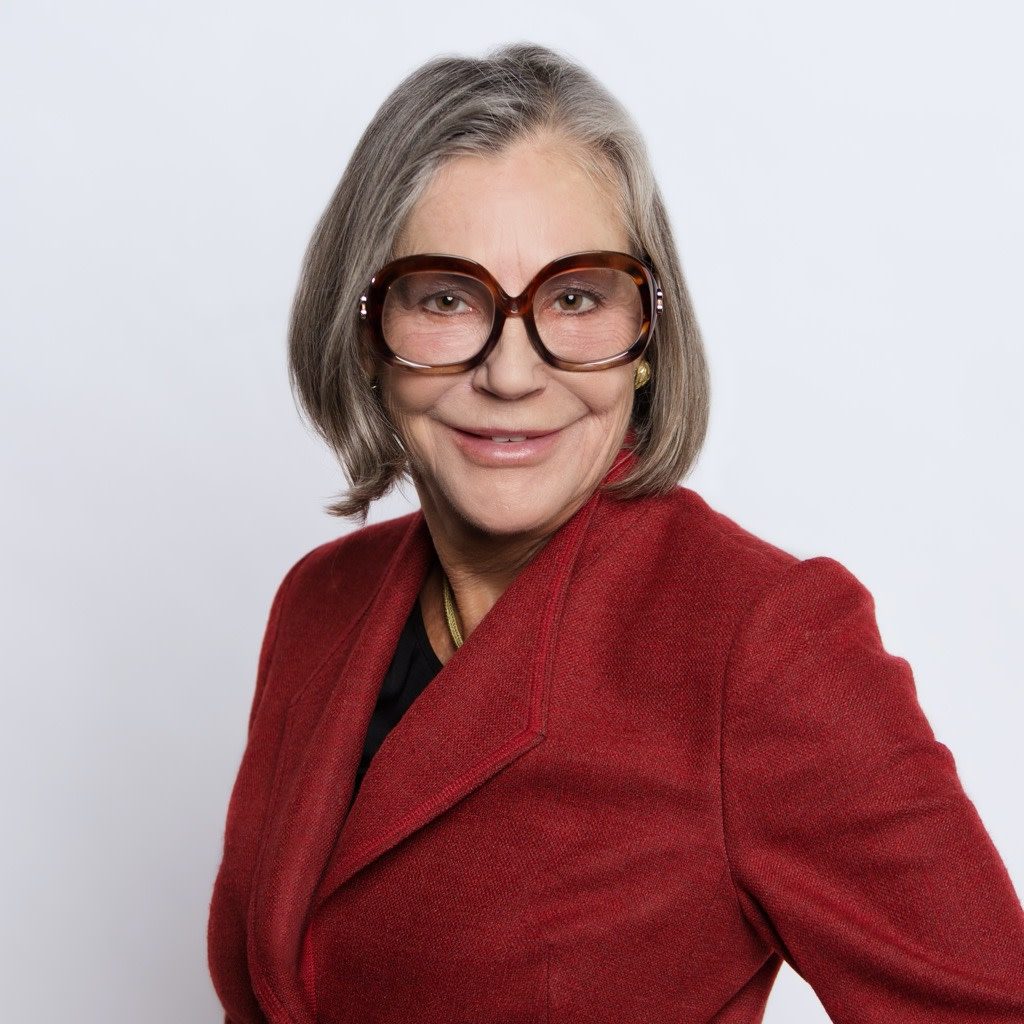 Older woman with gray hair, large brown glasses, red blazer, white background.