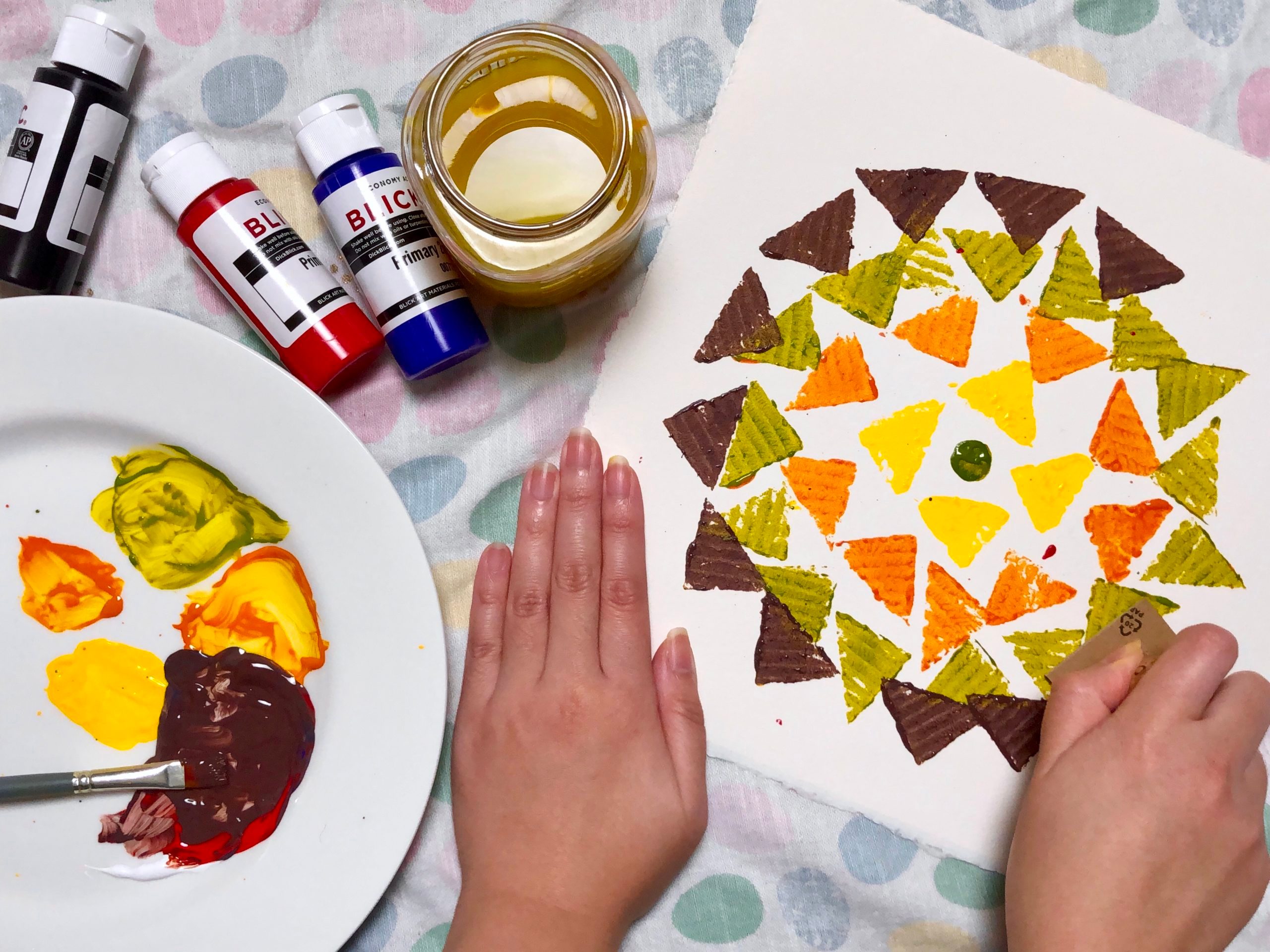 Mandala pattern with colored triangles and paintbrush on paper, next to paints and water jar.