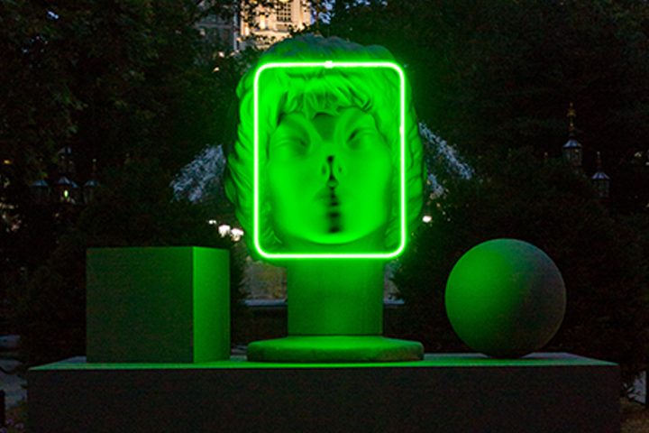 Glowing green neon outlines face sculpture with cube and sphere, dark outdoor backdrop.
