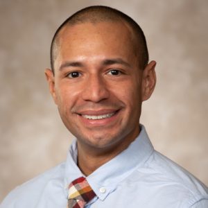 Smiling person in light blue shirt and multicolored tie on a neutral background.
