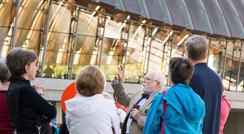 Tour guide gesturing to an attentive group outside a reflective modern building.