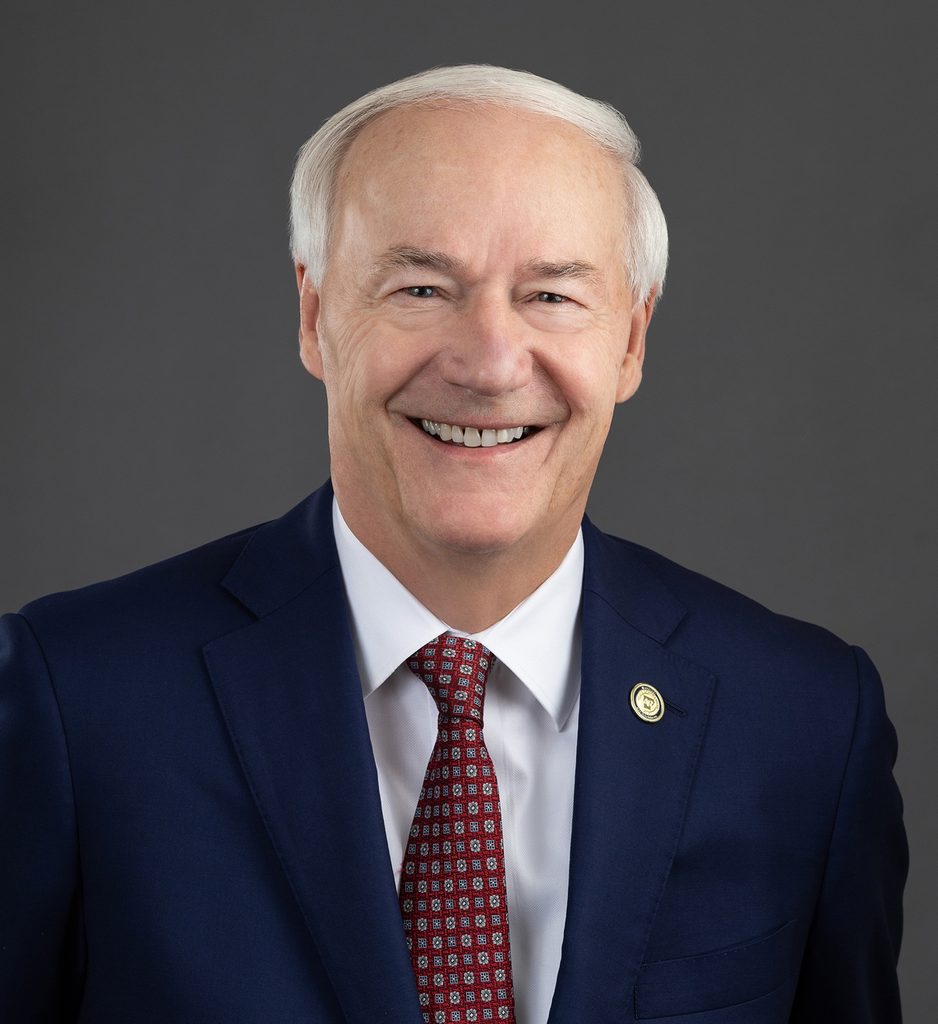 Older man with white hair in a blue suit, white shirt, and red patterned tie smiling on gray background.