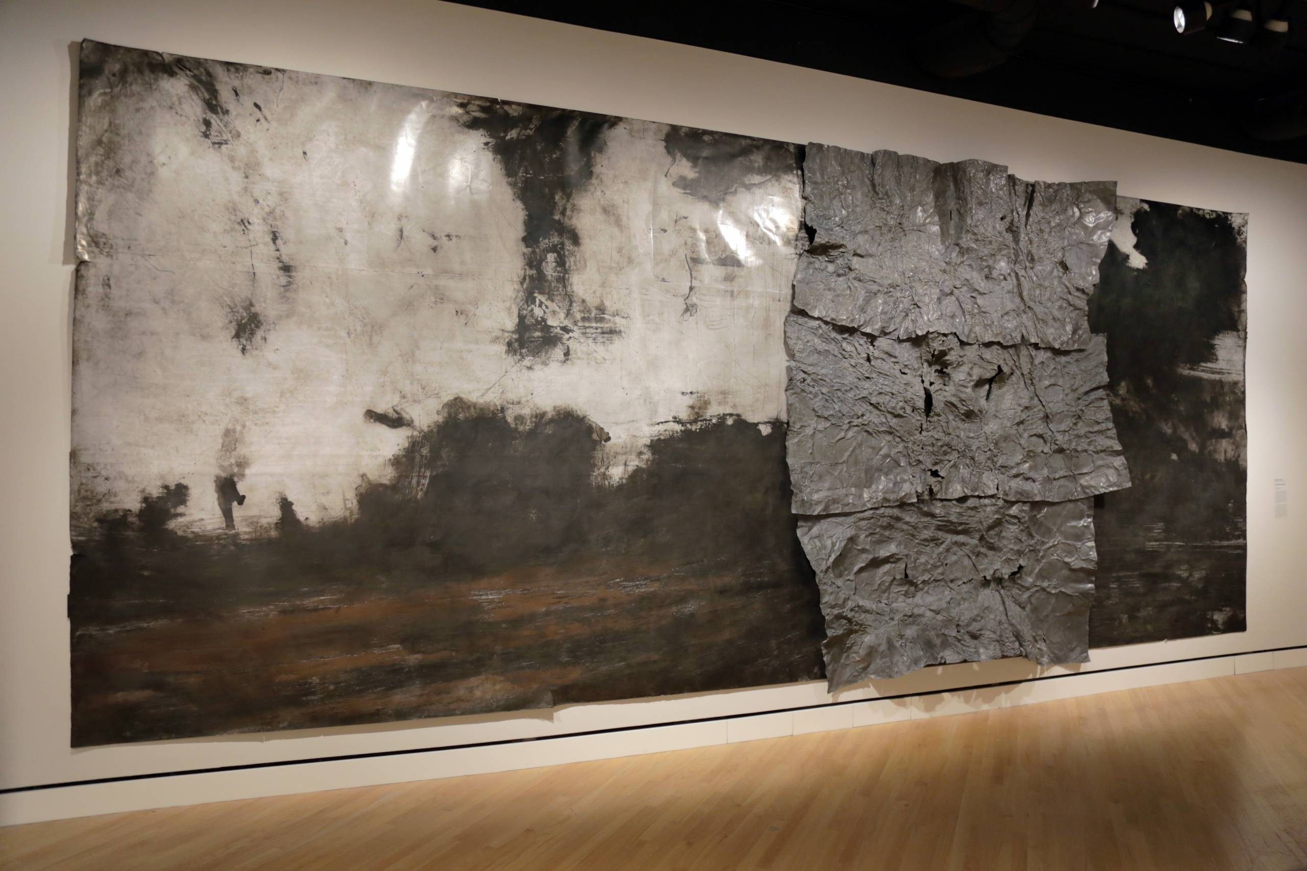 Abstract artwork on gallery wall with metallic sheets and dark cloud-like pattern.