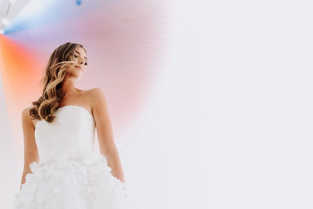 Woman in strapless white dress with wavy hair, colorful lighting, light background.