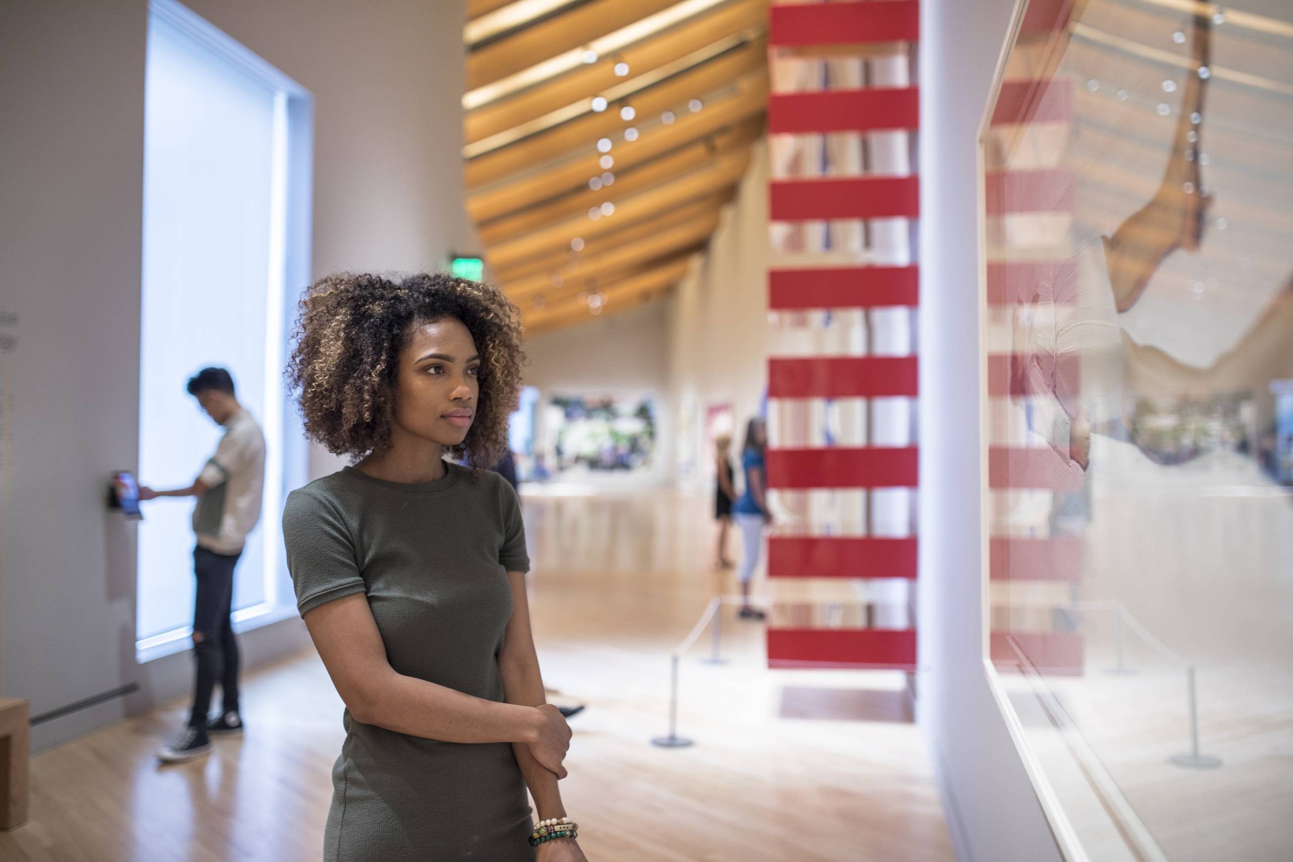 Woman viewing art in a gallery with a wooden ceiling and a man in the background.