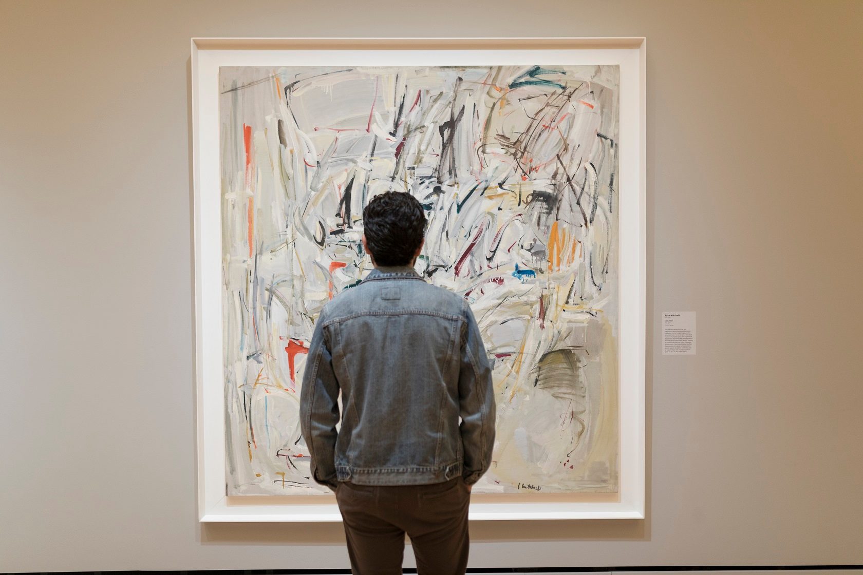 Person in denim jacket viewing an abstract painting with colorful brushstrokes in a gallery.