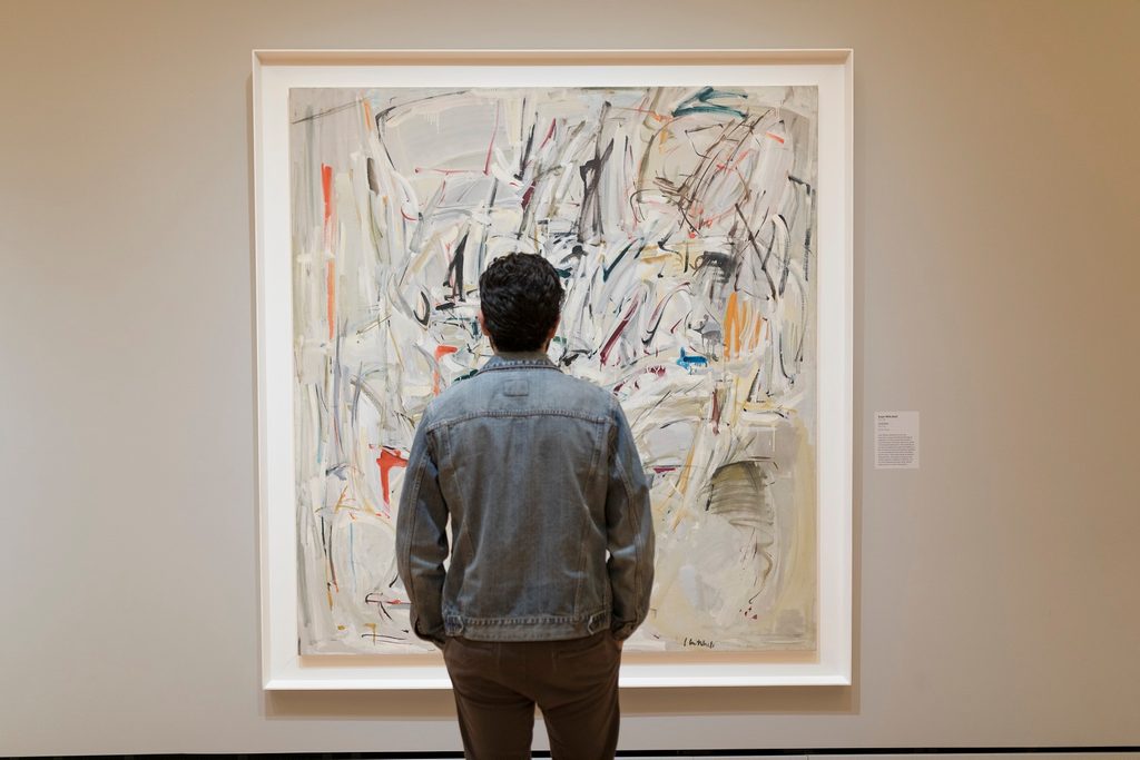 Person in denim jacket viewing an abstract painting with colorful brushstrokes in a gallery.