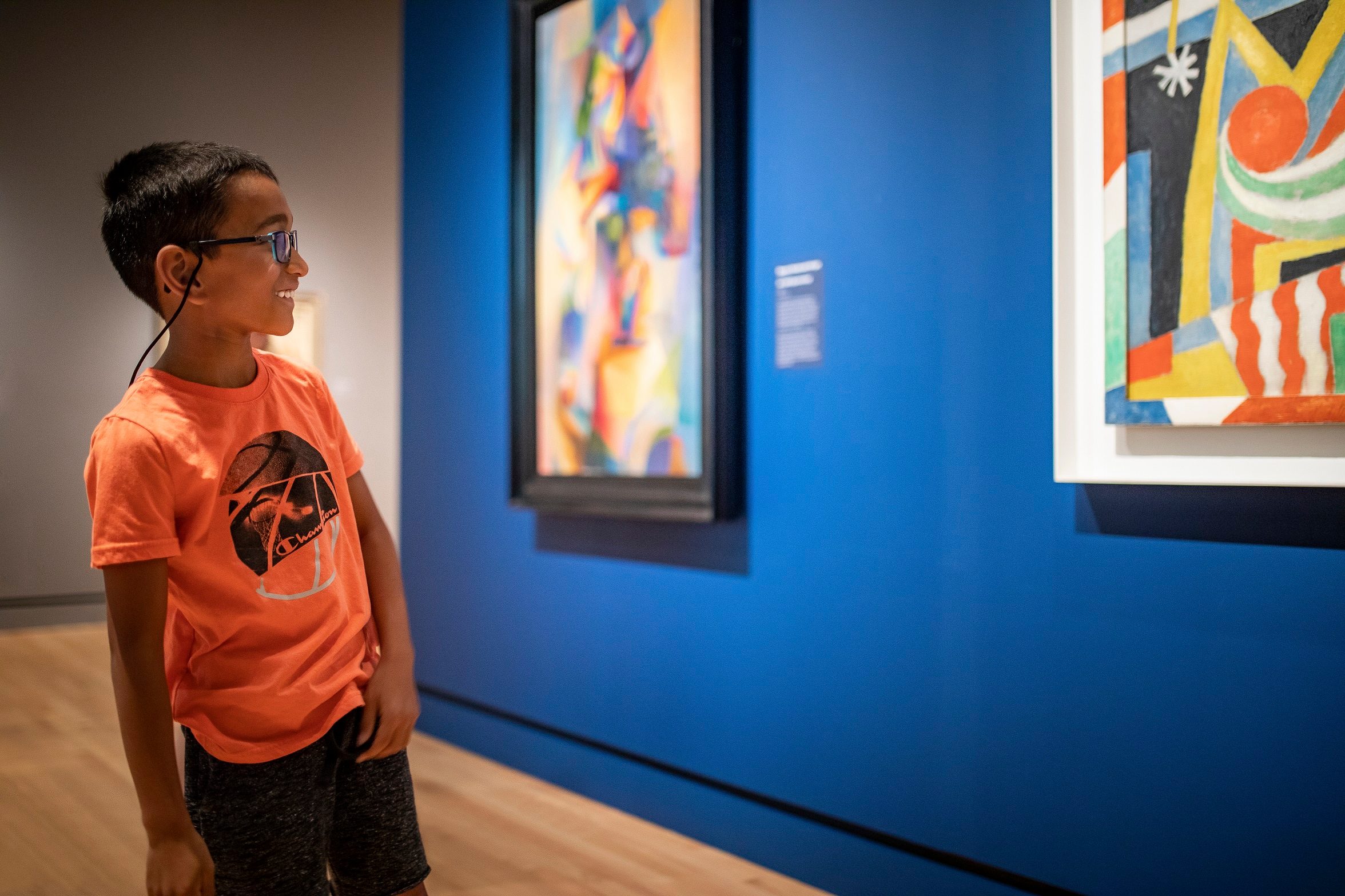Young boy in glasses and orange shirt viewing abstract paintings on a blue gallery wall.