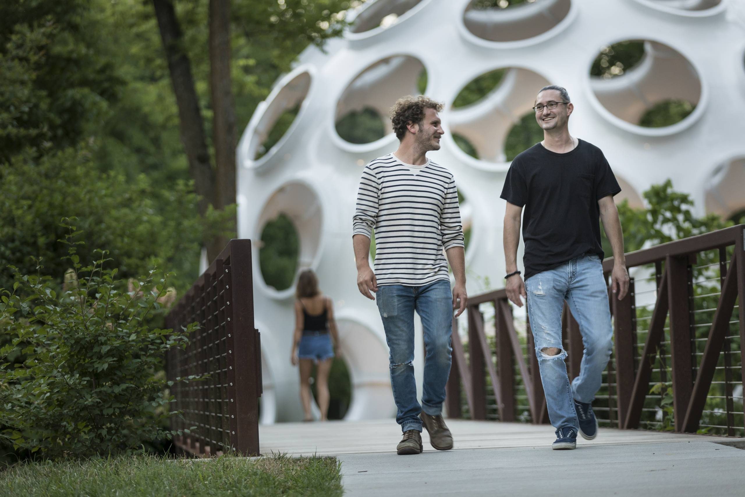 Two people walking on a bridge near greenery and a circular architectural structure.