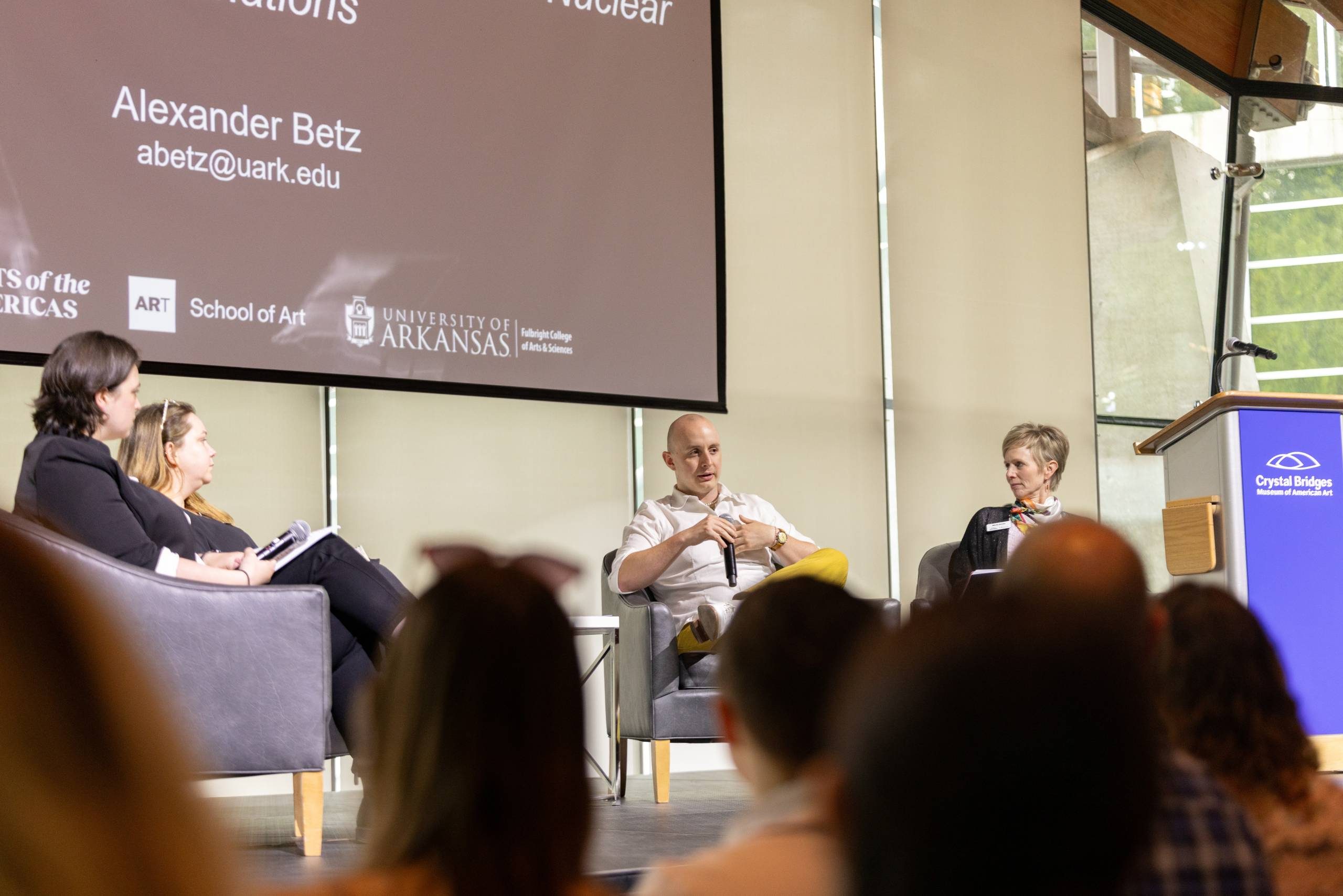 Panel discussion with three speakers on stage, University of Arkansas branding on screen.
