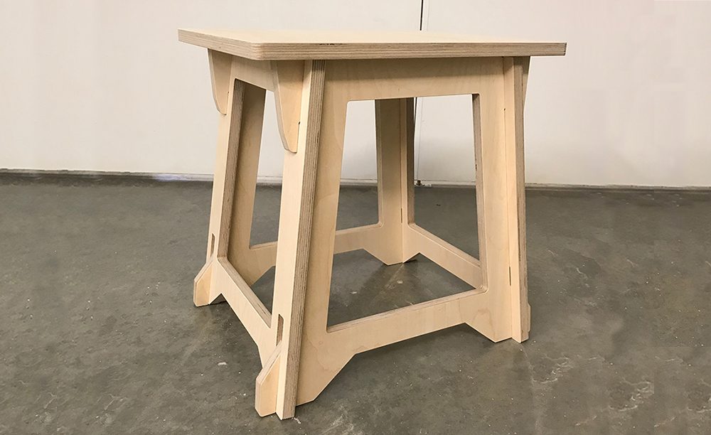 Wooden stool with square seat, interlocking legs, on concrete floor, plain wall background.