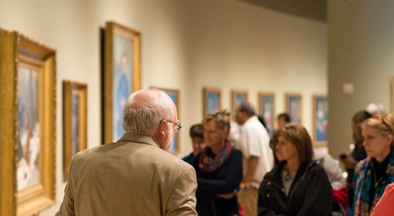 Group in art gallery viewing and discussing framed paintings on the walls.
