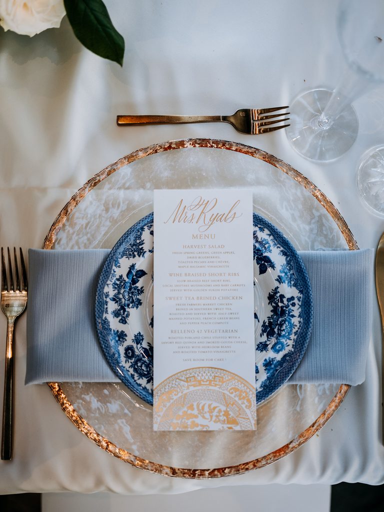White and blue floral plate on glass charger, gold rim, menu, fork, champagne flute, blue napkin.