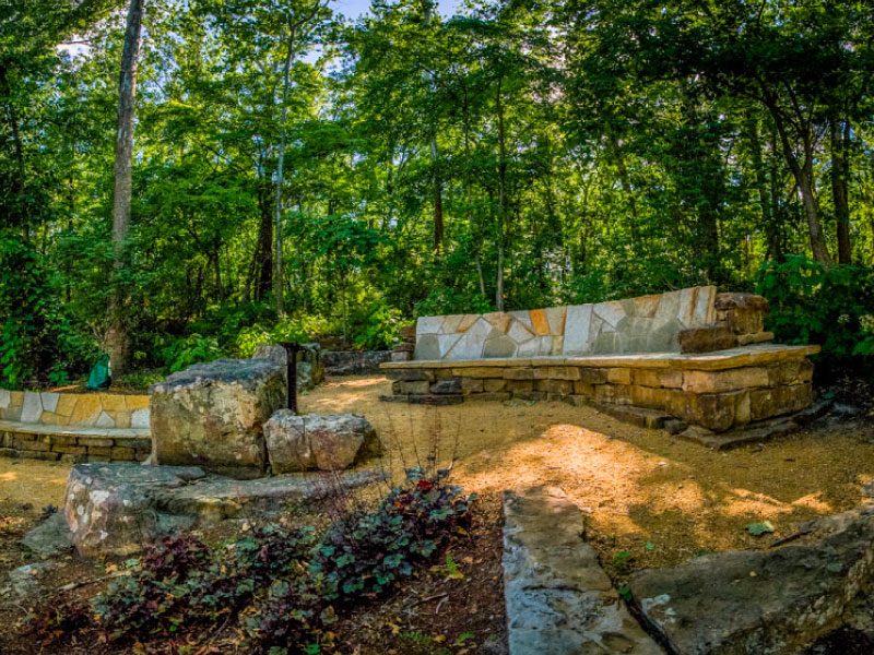 Outdoor stone seating area surrounded by trees and foliage, with a stone wall and dirt clearing.