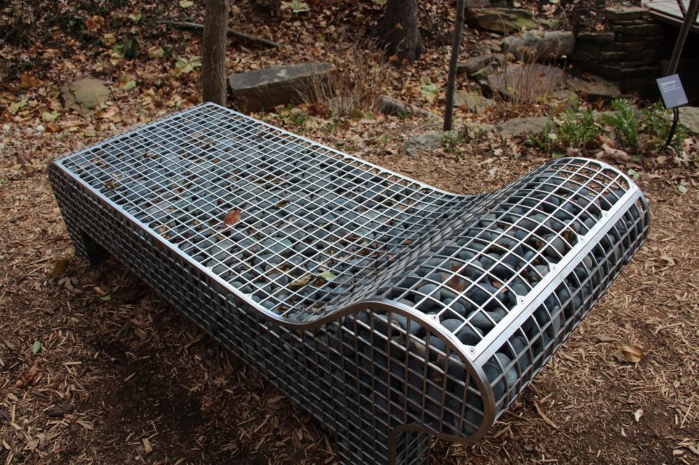 Outdoor bench with metal grid and stones in a natural setting with leaves and steps nearby.