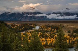 River winding through autumn forest with mountains and clouds in the background.