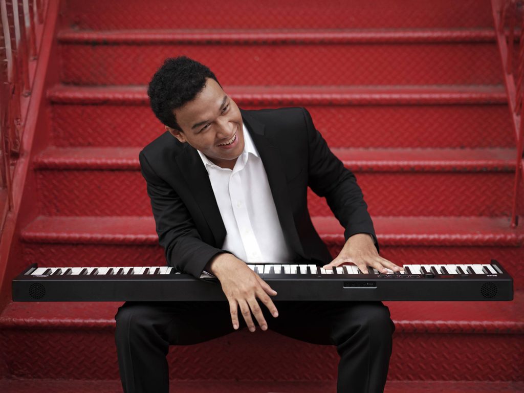 Person in black suit, sitting on red stairs with electronic keyboard, looking away and smiling.
