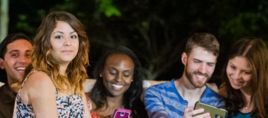 Five people sitting together, smiling at a phone, with greenery in the background.