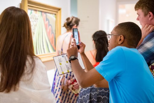 People viewing a painting in an art gallery; one taking a photo with a smartphone.