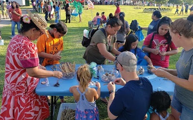 People at a blue table outdoors in a park, doing a group craft activity with materials.