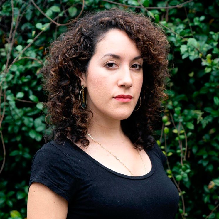 Woman with curly hair and hoop earrings, black top, leafy background.