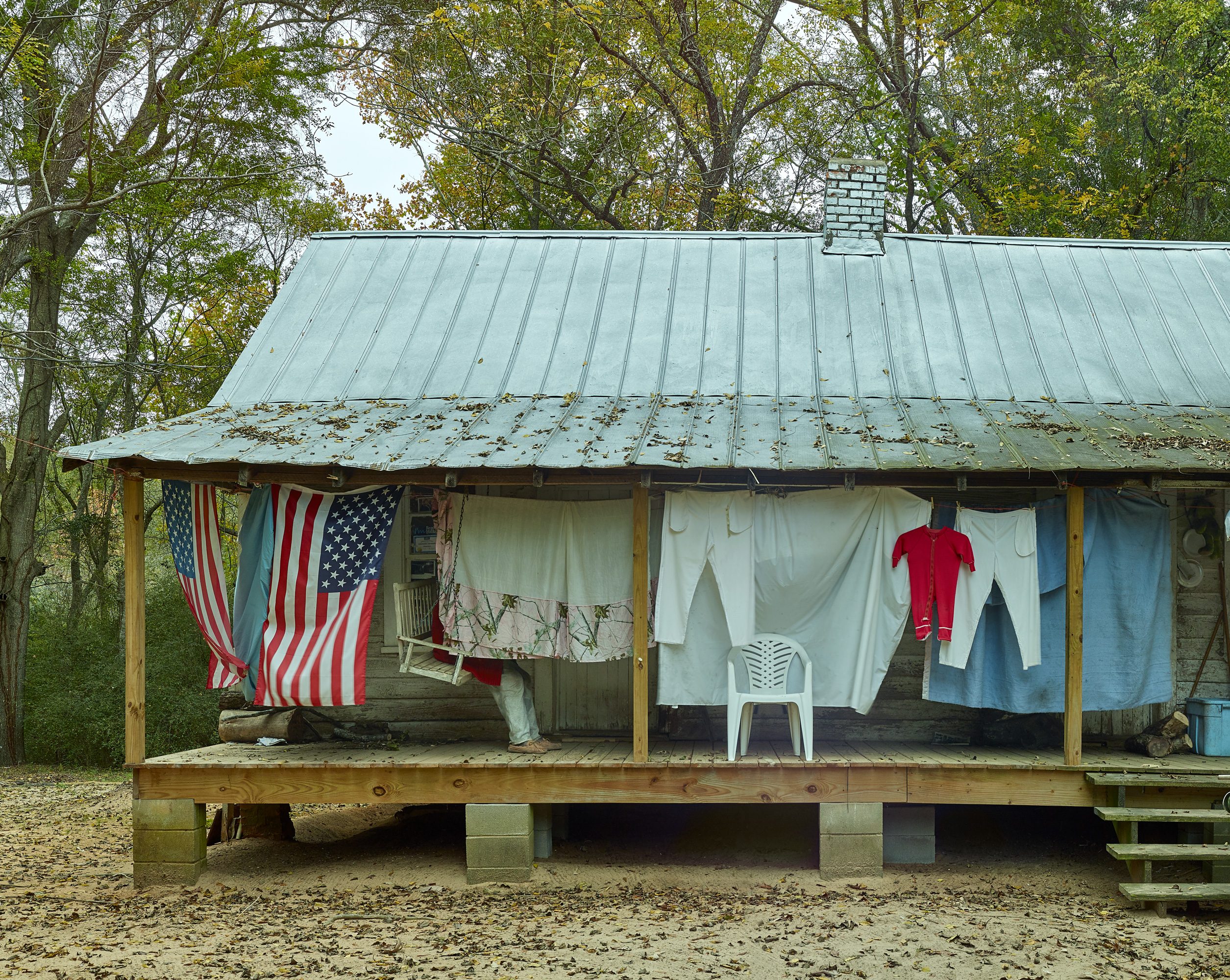 Rustic cabin with tin roof, porch, laundry, flags, and a white chair amid trees and fallen leaves.