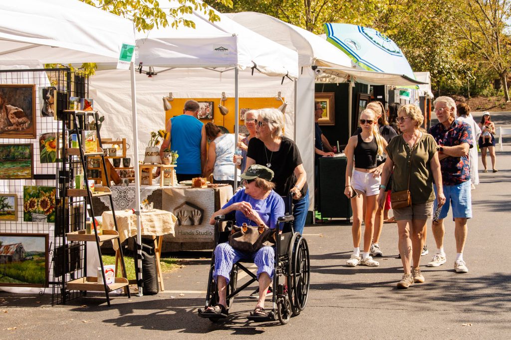 Outdoor art fair with people walking, browsing booths, and a woman pushing a wheelchair.