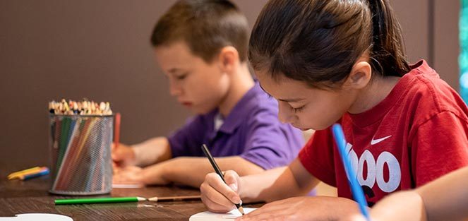 Children drawing at a table with colored pencils on paper; pencil container nearby.