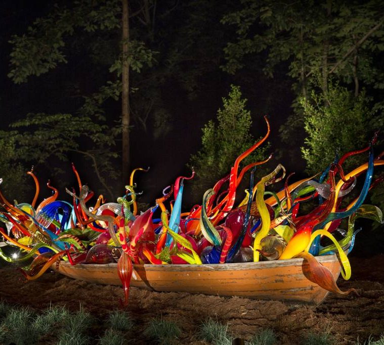 Old wooden boat with vibrant, colorful glass sculptures in natural setting at night.