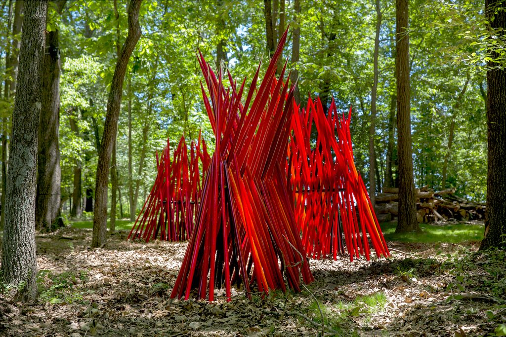 Red angular sculptures in a wooded area with trees and dappled sunlight.