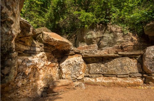 Natural rock formation with layered stones, lush greenery, and sandy ground.