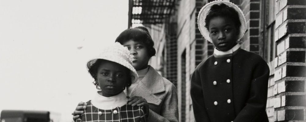 Three children stand solemnly on a sidewalk beside a brick building, wearing hats and coats.