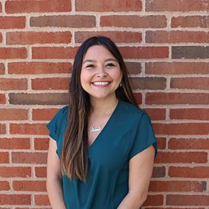 Person with long brown hair smiling in front of a brick wall wearing a teal top.