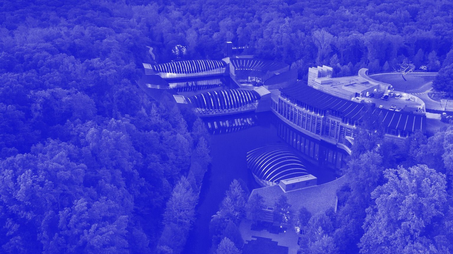 Modern architectural complex with curved roofs, connected by walkways over water, in a forest.