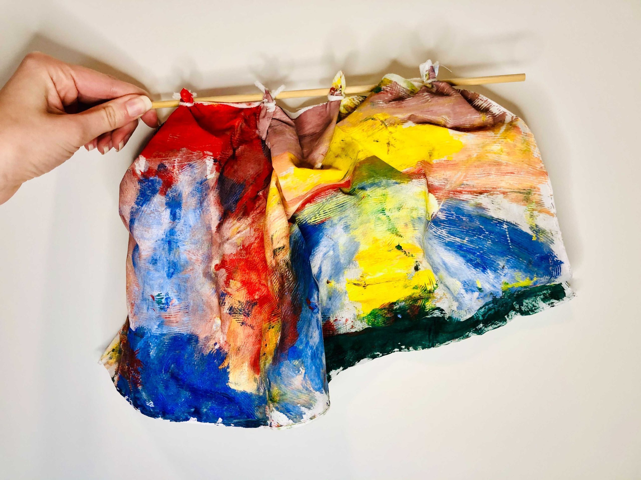 Hand holding a stick with colorful abstract brushstrokes on hanging paper.
