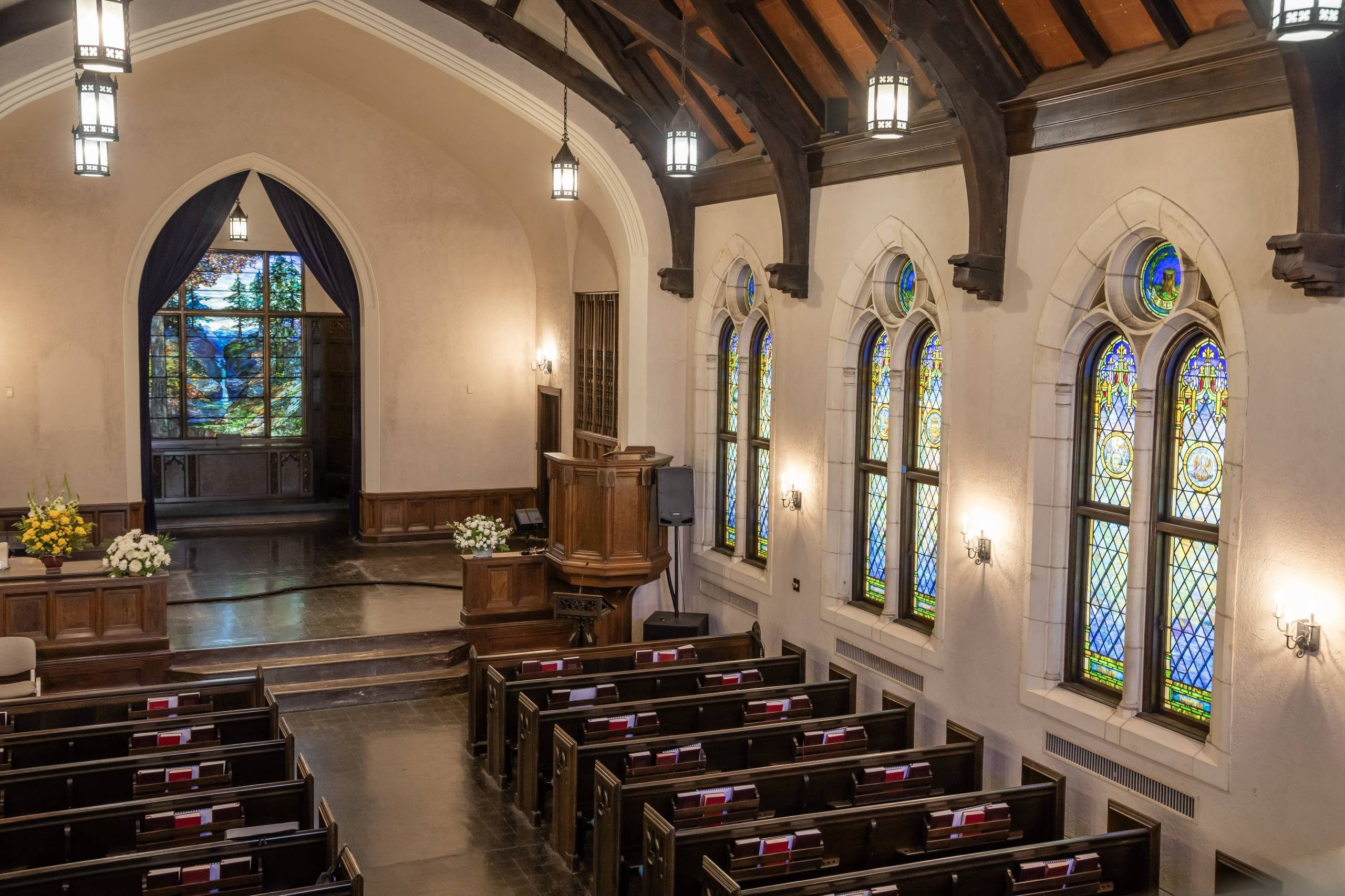 Church interior with wooden pews, pulpit, stained glass windows, lanterns, and floral arrangements.